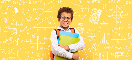 A young boy stands with a stack of books in his arms. He wears glasses and smiles broadly. Behind him is a bright yellow wall filled with math and science drawings and equations. © Prostock-studio