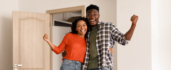 A couple stands in their new home, showing excitement and joy. They are smiling and cheering with arms raised, celebrating the moment. The room has light walls and an open door. © Prostock-studio