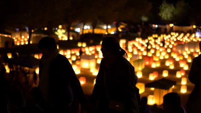 Panning shot of family silhouetted walking past rows of glowing paper lanterns at nighttime outdoor lantern festival