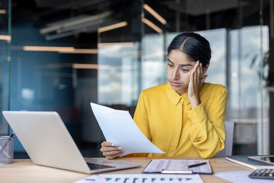 Upset and disappointed young Indian businesswoman sitting at the desk in the office and looking at the documents she is holding in her hand