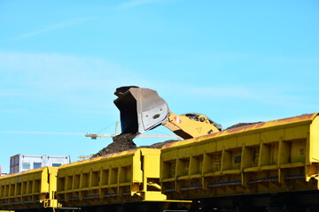 The bucket of a yellow excavator dumps dark earth into a railway gondola car on a sunny day © Wolf Steppe