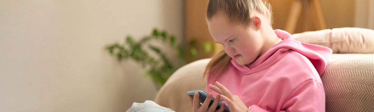 Teenage girl with Down syndrome using smartphone while sitting at home, engaging in digital communication and independent daily life. Ideal for inclusion, accessibility, social care campaigns, header