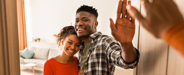 A smiling couple stands by the door, waving at a visitor. The man wears a plaid shirt while the woman is in a red top. The room has a light color scheme and a sofa in the background. © Prostock-studio