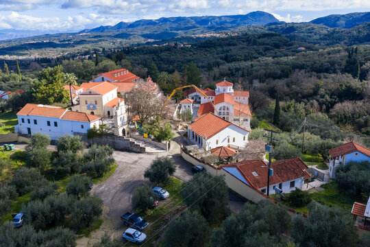 Wide aerial view of the historic Greek Orthodox Pantokrator Monastery in Agios Athanasios, Corfu. Traditional white buildings, red roofs, and green olive groves on a summer day.