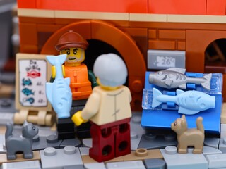 Obraz premium Tambov, Russian Federation - January 19, 2026 Lego fisherman minifigure selling a light blue fish from a fish stall to a customer on a city street. Two Lego cats standing near.