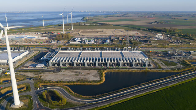 Aerial view of a large industrial data center facility and wind turbines along the coast under a clear sky in Eemshaven, Groningen, Netherlands.