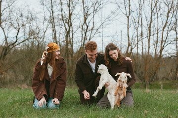Fototapeta premium Young man and two women enjoy a spring day outdoors while playing with two baby goats in a grassy field surrounded by bare trees under an overcast sky