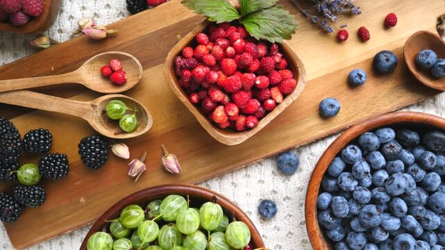 Colorful assortment of fresh summer berries on a rustic table with wooden bowls and spoons, featuring wild strawberries, blueberries, gooseberries, blackberries, and raspberries