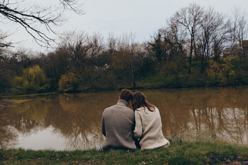 Obraz premium A couple sits close by a calm lake in autumn, wearing cozy sweaters, sharing a quiet moment, soft breeze, muted colors, reflective water, peaceful countryside setting
