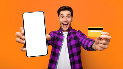 A man smiles widely while holding a large smartphone in one hand and a credit card in the other. He stands in front of a bright orange backdrop, looking happy and engaged. © Prostock-studio
