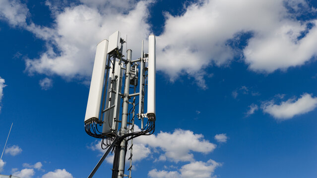 Aerial view of a telecommunications tower with white panel antennas against a bright blue sky with scattered white clouds in Amsterdam, North Holland, Netherlands.