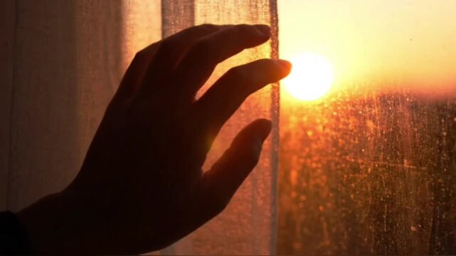 Silhouette of female hand slowly touching window glass at beautiful sunset or sunrise with bright sun disc on horizon, inspiring and spiritual moment.