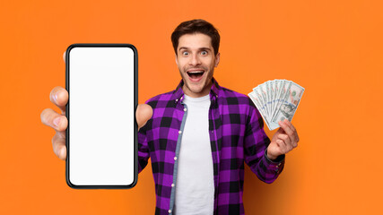A man stands with a bright orange background, holding a large smartphone in one hand and a bundle of cash in the other. He shows a big smile that expresses excitement and joy. © Prostock-studio