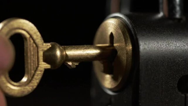 Macro closeup of vintage brass key slowly inserted into dark metal padlock keyhole mechanism to unlock security device for opening wooden door during home visit.