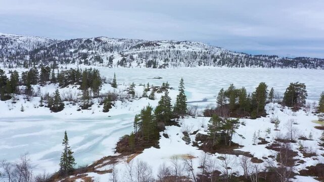 Aerial forward motion over snow-covered landscape at Hallbjonnsekken by Dalen , showcasing frozen lake surrounded by evergreen trees and distant mountains in spring tranzition under moody spring sky. 