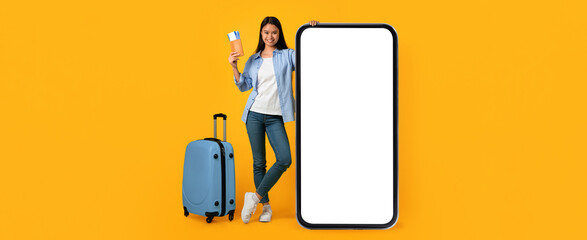 Woman holds travel tickets and stands next to a large blank screen. She leans against the screen while a blue suitcase rests nearby. The background is bright yellow. © Prostock-studio