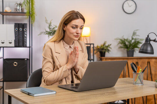 Middle-aged businesswoman at home office folds hands in prayer at laptop looks up hopes for approval. Freelancer girl at table waits for reply checks inbox breathes writes note stays calm faith inside