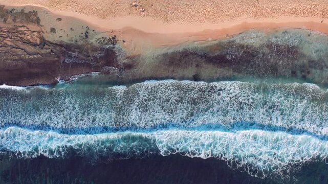 Aerial top down view of a Hawaiian coastline where waves crash over shallow reef formations, creating intricate vein‑like patterns beneath the water. The turquoise and deep‑blue ocean transitions into
