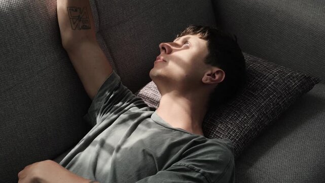Young man resting on sofa with sunlight on face, touching light with hand and enjoying quiet moment, anti productivity, mindfulness and emotional recovery concept