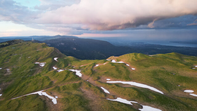 Aerial view of Le Colomby de Gex mountain ridge with green slopes and patches of snow under dramatic storm clouds at sunset in Echenevex, Auvergne-Rhone-Alpes, France.