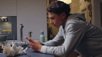 Fototapeta na wymiar Teenager leaning over kitchen counter using smartphone with focused expression, absorbed in quiet digital moment inside home environment, conveying modern contemplative lifestyle