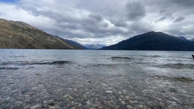 Lago Puelo National Park in Patagonia, Argentina, with gentle waves rolling toward the camera beneath a dramatic overcast sky. Cinematic lake scene featuring a distant horizon, dark clouds.