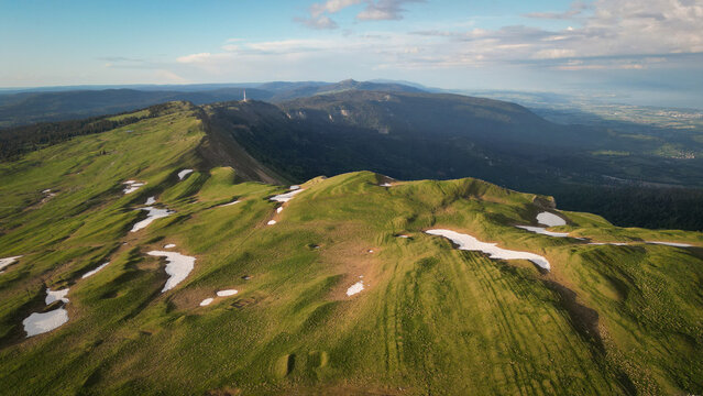 Aerial view of Le Colomby de Gex mountain ridge with green grassy slopes and melting snow patches in the Jura Mountains Echenevex, Auvergne-Rhone-Alpes, France.