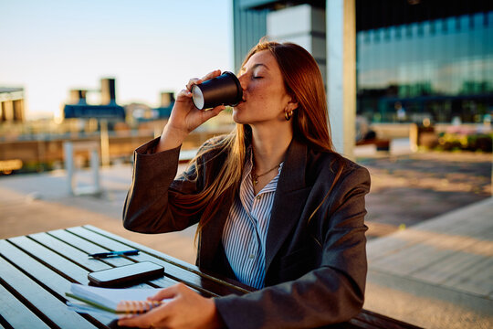 Young businesswoman with freckles drinking coffee from a disposable cup outdoors during golden hour, relaxing at a wooden table with a notebook and smartphone