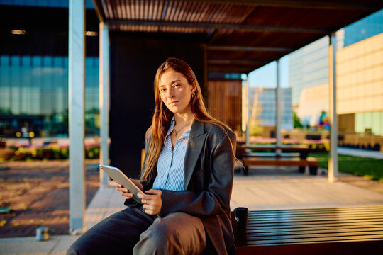 Young professional businesswoman sitting on a bench in a modern urban park, holding a digital tablet and looking directly at the camera with a confident expression during sunset
