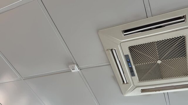 Ceiling cassette air conditioner unit mounted in suspended office ceiling grid, white grille and vents, closeup inspection