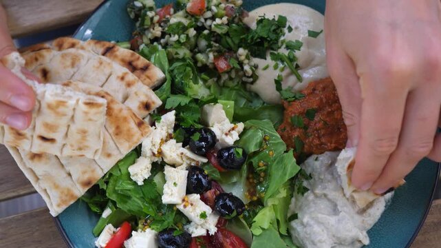 Close up of female hands tearing a piece of warm pita bread and dipping it into baba ghanoush on a mediterranean mezze platter with hummus, tabbouleh, and feta cheese salad