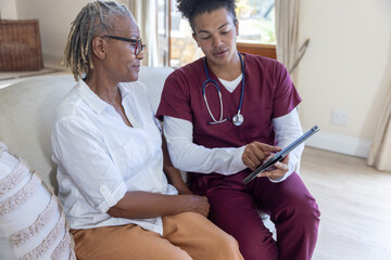 Fototapeta premium Mid-adult nurse in scrubs with stethoscope on sofa pointing tablet to senior African American woman