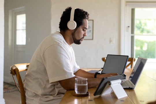 Mid-adult African American man wearing headphones, working on two laptops, tablet in dining area