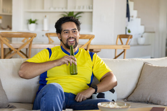 African American man watching TV on sofa, wearing jersey, holding green bottle, soccer ball nearby