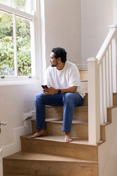African American man sitting barefoot on wooden stairs holding smartphone, looking out large window