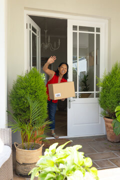 Senior Asian woman standing in white glass door frame holding labeled cardboard package and waving