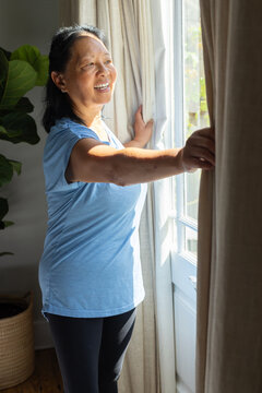 Senior Chinese woman pulling beige curtains at glass door at home, wearing blue shirt and leggings