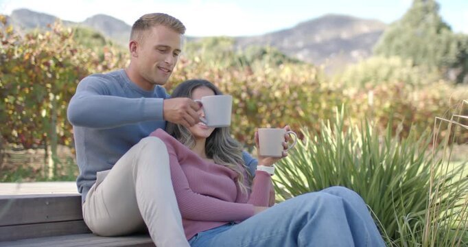 Couple cuddling on steps, man leaning in offering mug, sipping alternately taking in vineyard view