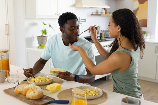 Couple sharing breakfast at kitchen island, woman in green top feeding man spoonful, eggs and bread
