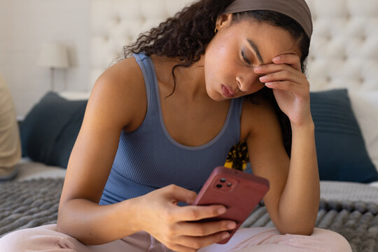 African American woman in her twenties sitting on bed wearing blue tank top holding pink smartphone