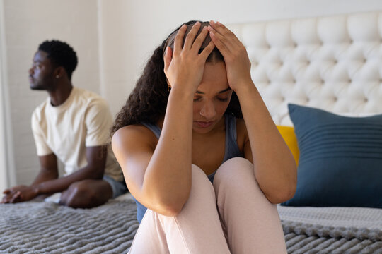 African American couple sitting on gray bedspread in bedroom, woman pressing head, man turning away