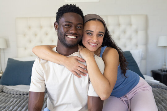Diverse couple smiling, woman wrapping arms around man sitting on bed with headboard and lamps