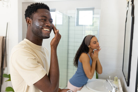 African American couple smiling while applying facial cream at bathroom vanity with jar