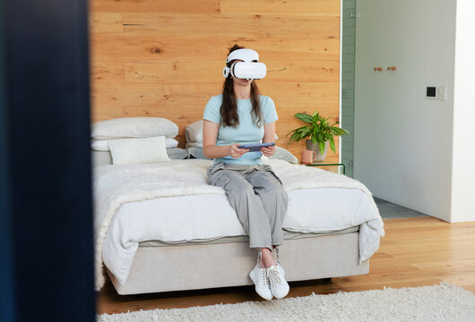 Woman wearing white VR headset sitting on edge of bed holding controller in bedroom