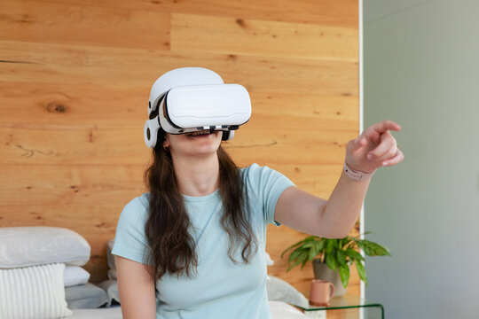 Woman in her 20s wearing white VR headset, reaching forward on bed near wooden wall