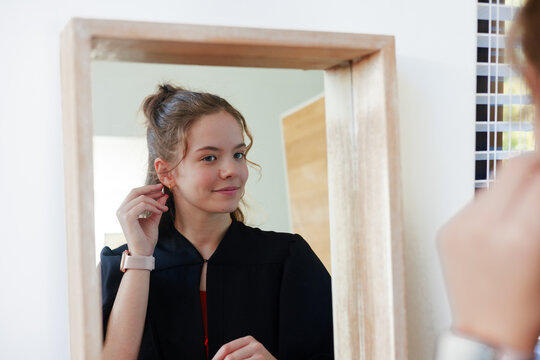 Woman fixing earring while looking into wood frame mirror at home wearing dark robe with smartwatch