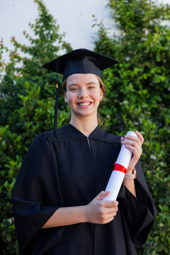 Female graduate standing in front of hedge in cap and gown holding diploma with red ribbon
