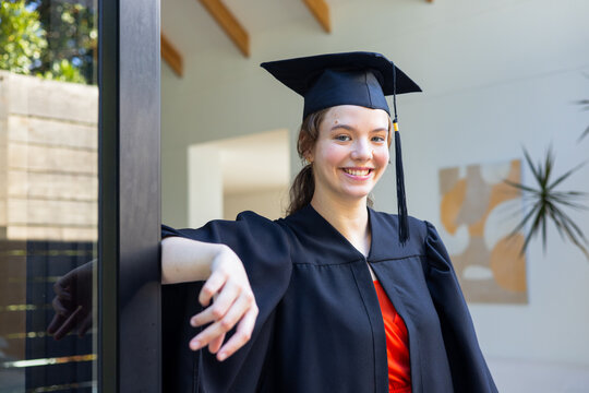 Female graduate wearing black gown and mortarboard, leaning on glass doorframe at home with red top