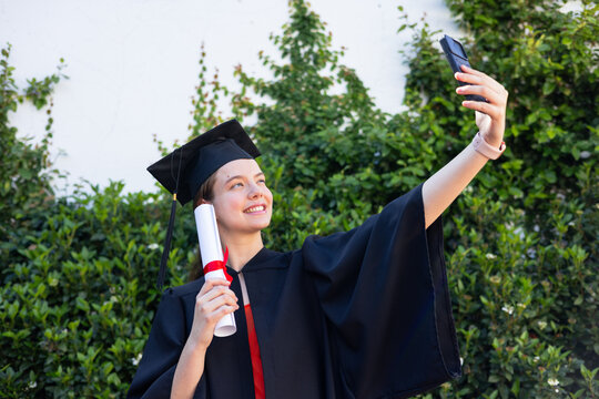 Female graduate wearing gown and mortarboard holding diploma and taking smartphone selfie by hedge