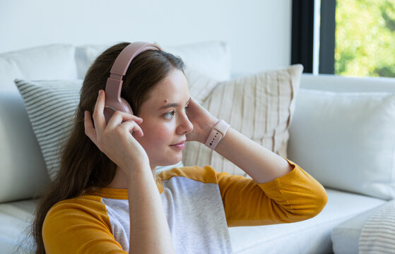 Woman in twenties in mustard raglan sitting at home adjusting pink headphones, smartwatch visible